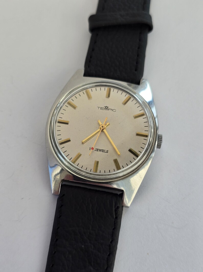 May include: A vintage TEMPIC wristwatch with a silver case and a cream face. The watch has gold hands and markers, with the text "TEMPIC" and "1 Jewels". The watch has a black leather strap.