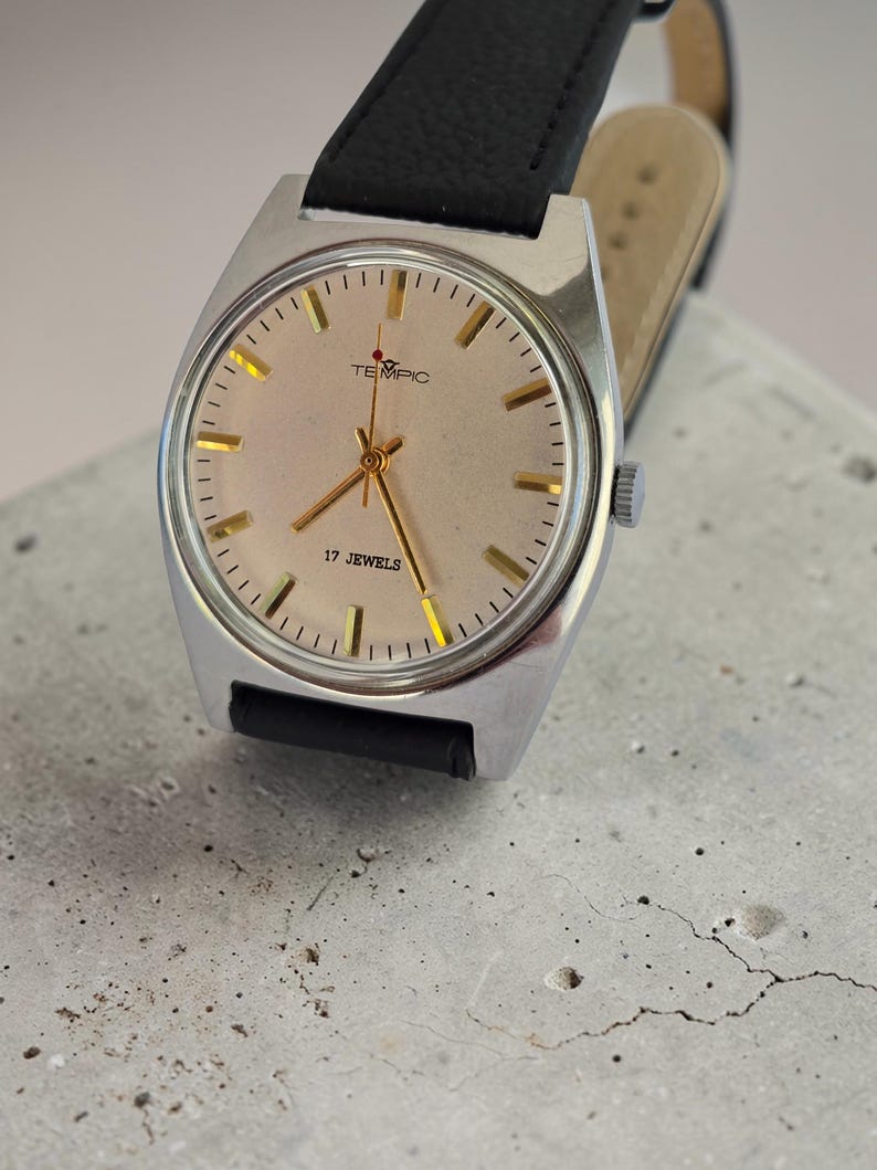 May include: A vintage wristwatch with a silver case and a light beige face. The watch has gold hands and markers, with the text "TEMPIC" and "17 JEWELS". It features a black leather strap.