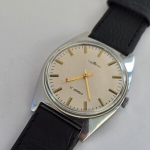 May include: A vintage TEMPIC wristwatch with a silver case and a light-coloured face. The watch has gold hands and markers, with the text "17 JEWELS" and the brand name "TEMPIC". It features a black leather strap.