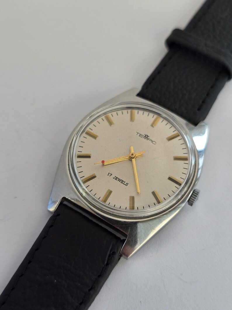 May include: A vintage TEMPIC wristwatch with a silver case and a light-coloured face. The watch has gold hands and markers, with the text "17 JEWELS" and the brand name "TEMPIC". It features a black leather strap.