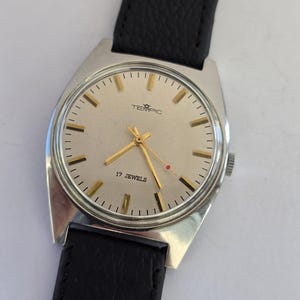 May include: A vintage TEMPIC wristwatch with a silver case and a light-coloured face. The watch has gold hands and markers, a black leather strap, and the text "17 JEWELS".