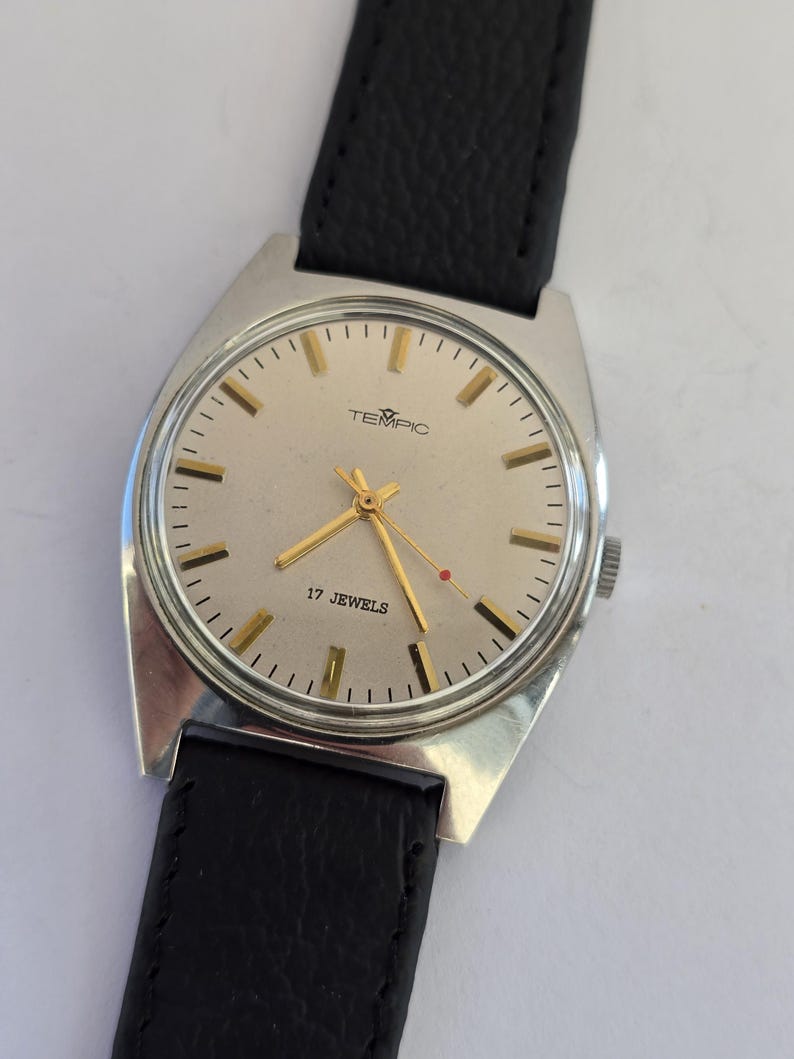 May include: A vintage TEMPIC wristwatch with a silver case and a light-coloured face. The watch has gold hands and markers, a black leather strap, and the text "17 JEWELS".