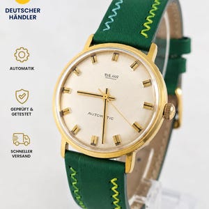 May include: A gold-toned wristwatch with a cream-colored face and gold hands and markers. The watch has a green leather band with decorative stitching. The watch face reads "BEHA" and "AUTOMATIC".