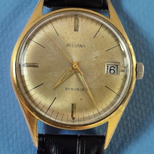 May include: A vintage gold-toned wristwatch with a black leather band. The watch face is a light gold color with black lines and the word "BELLANA" printed on it. The watch also has a date display showing the number "16".