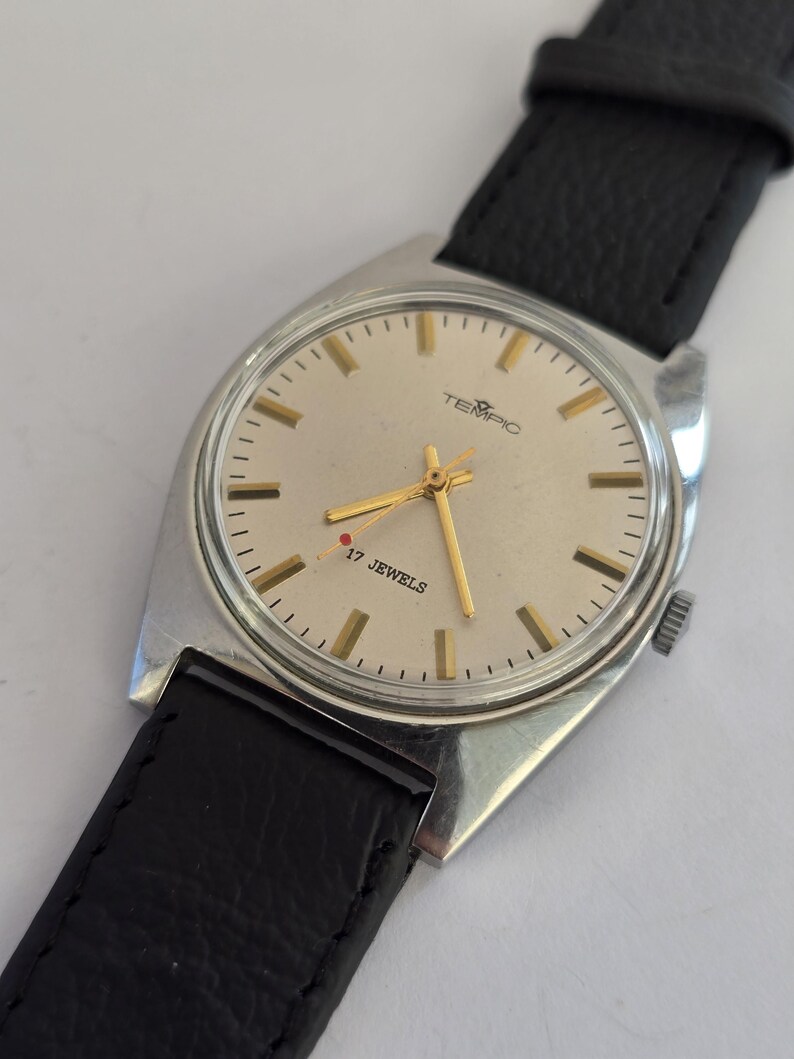 May include: A vintage TEMPIC wristwatch with a silver case and a black leather strap. The watch face is light-coloured with gold hands and markers. The text "TEMPIC" and "17 JEWELS" are visible on the watch face.