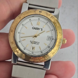 May include: A silver and gold-toned Gaddy's watch with a white face, gold hands, and a date display. The watch features a tachymeter bezel and a stainless steel band. The watch face displays the text "AUTOMATIC INCABLOC".