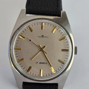 May include: A silver TEMPIC wristwatch with a light-coloured face, gold hands and markers, and a black leather strap. The watch face displays the text "TEMPIC" and "17 JEWELS".