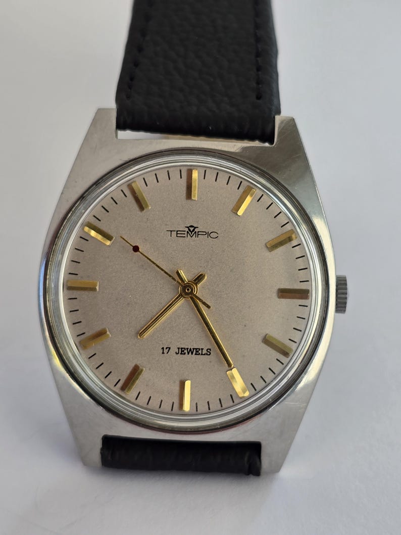 May include: A silver TEMPIC wristwatch with a light-coloured face, gold hands and markers, and a black leather strap. The watch face displays the text "TEMPIC" and "17 JEWELS".