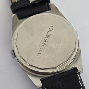 May include: Close-up of the back of a silver watch with a black leather strap. The watch face has the text "TEMPIC01" printed on it. The watch band is black leather with a light tan underside.