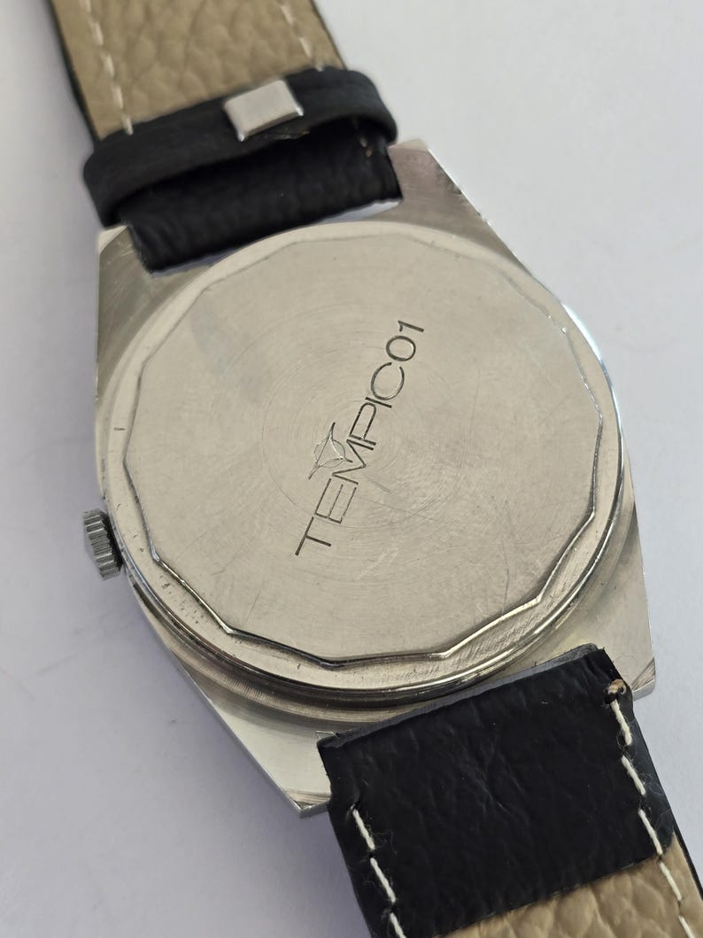 May include: Close-up of the back of a silver watch with a black leather strap. The watch face has the text "TEMPIC01" printed on it. The watch band is black leather with a light tan underside.