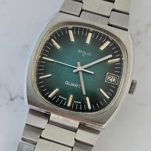 Könnte beinhalten: Eine Vintage-Armbanduhr in Silber mit quadratischem Zifferblatt und grünem Zifferblatt. Das Zifferblatt zeigt den Text "APOLLO 10 QUARTZ" und das Datum "20". Die Uhr hat ein silbernes Armband.