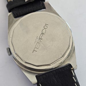 May include: Close-up of a silver watch back with a black leather strap. The watch face has the word "TEMPICO1" printed on it. The watch has a rectangular shape and a silver bezel.