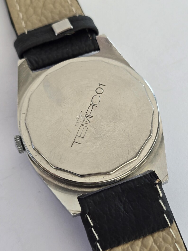 May include: Close-up of a silver watch back with a black leather strap. The watch face has the word "TEMPICO1" printed on it. The watch has a rectangular shape and a silver bezel.