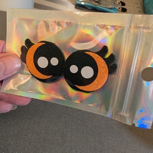 Puede incluir: Dos adornos de fieltro negro con temática de Halloween, con lunas crecientes naranjas y ojos blancos, empaquetados en una bolsa holográfica iridiscente. Para manualidades o scrapbooking.