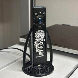 Puede incluir: Linterna negra Olight M1T con un diseño de dragón blanco y el logotipo de Olight. La linterna está en un soporte negro impreso en 3D. El soporte tiene una base de panal y soportes curvos. La linterna tiene una lente azul.