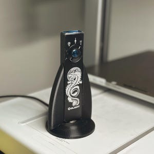 May include: Black Olight flashlight with a white dragon design and the Olight logo. The flashlight is mounted on a black stand. The stand is on a white surface.