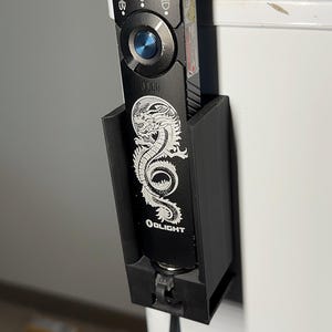 Può includere: Torcia Olight nera con un design a drago, tenuta in un supporto nero stampato in 3D. La torcia ha un pulsante blu e vari simboli. Il supporto è montato su una superficie bianca.