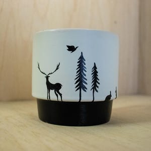 Könnte beinhalten: Zylindrischer Keramik-Pflanztopf mit schwarzem Sockel und weißem Oberteil. Der weiße Teil zeigt eine schwarze Waldszene mit einem Hirsch, Bäumen, einem Vogel und einem Kaninchen. Geeignet für kleine Pflanzen.