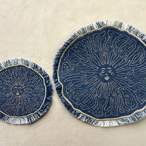 May include: Two circular denim fabric mats with frayed edges, featuring a gold embroidered sun face design with radiating rays. The smaller mat is on the left and the larger mat is on the right, both showcasing a dark blue base with metallic gold stitching.