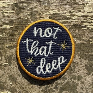 May include: A circular navy blue embroidered patch with the words "not that deep" in white script. The patch has a gold border and two small gold starburst designs. The patch is on a weathered wooden surface.