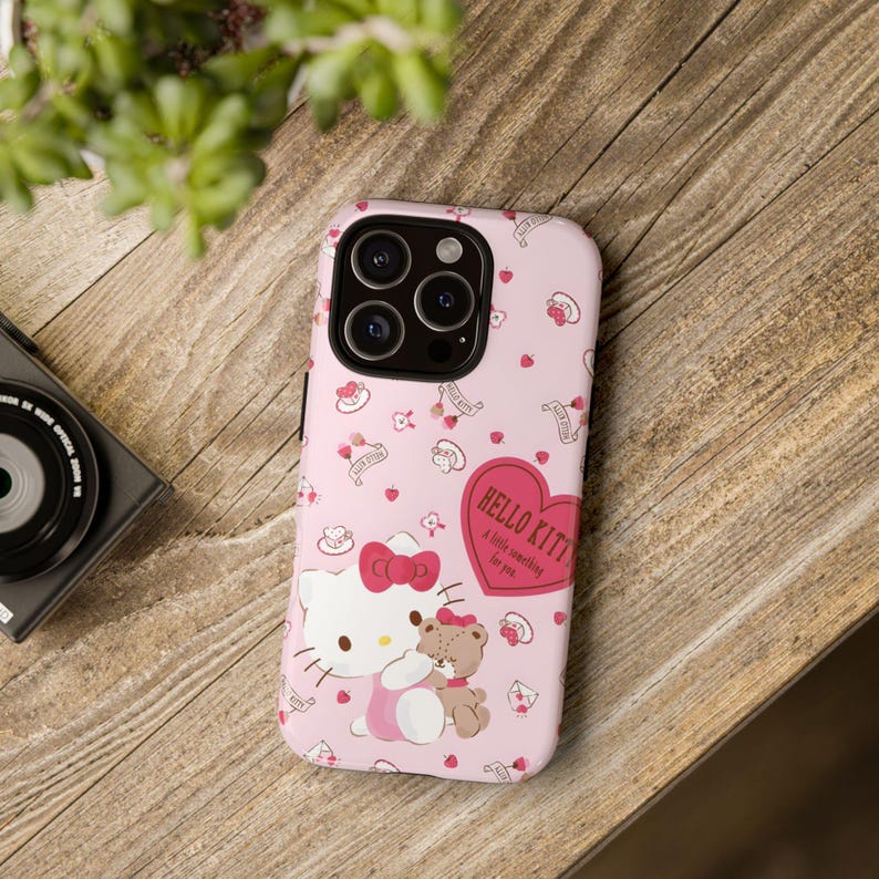 May include: Pink phone case featuring Hello Kitty with a teddy bear, surrounded by hearts and small desserts. The case has the text "Hello Kitty, a little something for you."