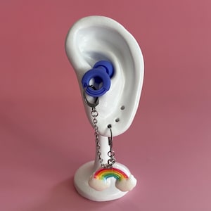 Puede incluir: Un pendiente azul con una cadena y un dije de arcoíris. El dije de arcoíris tiene colores rojo, naranja, amarillo, verde y azul. El pendiente se muestra en un soporte blanco en forma de oreja sobre un fondo rosa.