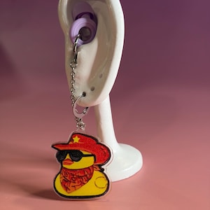 May include: A silver-toned metal earring featuring a dangling acrylic charm of a yellow rubber duck wearing a red cowboy hat with a yellow star, black sunglasses, and a red patterned bandana, attached to a small silver chain and hoop.