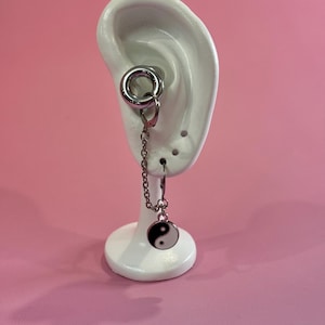 Peut inclure: Une boucle d'oreille argentée avec une chaîne et un charme pendant en forme de symbole yin-yang noir et blanc. La boucle d'oreille est présentée sur un modèle d'oreille blanc sur fond rose.