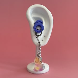 May include: A white ear-shaped display stand holds blue earplugs and a dangling earring. The earring has a small, colourful cat charm with yellow, pink, and blue hues. The background is a solid pink.