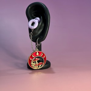 Può includere: Un orecchino a cerchio color argento con un ciondolo circolare pendente su sfondo smaltato rosso. Il ciondolo raffigura un gruppo di funghi e una serie di fasi lunari lungo il bordo superiore. Una sottile catenella collega il ciondolo all'anello.