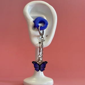 Peut inclure: Boucle d'oreille papillon bleue avec une chaîne argentée et une jauge d'oreille bleue, présentée sur un support blanc en forme d'oreille. Le papillon a un corps et des ailes bleu foncé avec un contour argenté. Le fond est rose uni.