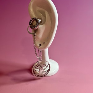 Peut inclure: Une boucle d'oreille en argent avec une chaîne et un pendentif circulaire. Le pendentif présente un motif découpé d'un soleil, de nuages et d'un OVNI. La jauge est insérée dans un modèle d'oreille blanc, sur fond rose.