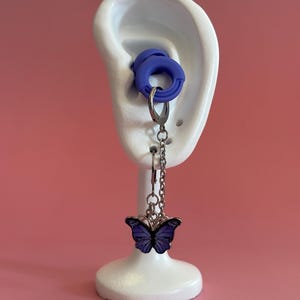 Puede incluir: Pendientes con un dije de mariposa. La mariposa es de color púrpura oscuro con detalles negros, suspendida de una cadena plateada. Los pendientes se exhiben en un soporte blanco con forma de oreja, con un tapón para los oídos azul.