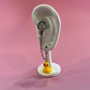 May include: A silver ear gauge with a chain and a small yellow rubber duck charm. The duck has an orange beak and is attached to a small hoop earring. The gauge is displayed on a white ear-shaped stand against a pink background.