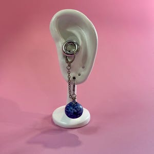 May include: A silver earring featuring a chain and a dark blue, speckled, round bead. The earring is shown on a white ear-shaped stand against a pink backdrop. The earring is a dangle style.