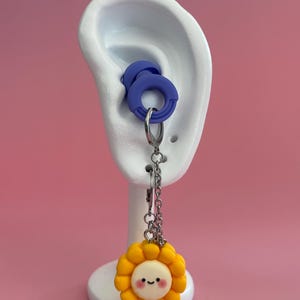 May include: A white ear model displays blue ear gauges. A silver chain hangs from the gauge, supporting a yellow and white sunflower charm with a smiling face. The background is a solid pink.