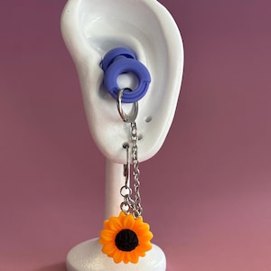 May include: Earrings with a sunflower charm. The earrings have a silver chain and a bright orange and black sunflower charm. The earrings are displayed on a white ear-shaped stand, with a blue ear plug in the ear.
