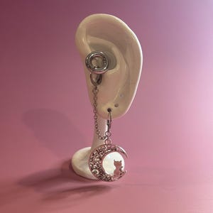May include: A silver earring with a crescent moon pendant. The moon has a filigree design and a cat silhouette. The earring is attached to a silver tunnel. The earring is displayed on a white ear model.