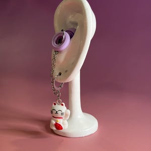 May include: A silver-toned chain earring featuring a white and red beckoning cat charm. The earring includes a light purple circular component that sits within the ear, connected by a metal chain to a small hoop earring at the bottom lobe.