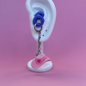 May include: Blue ear gauges and a pink cowboy hat earring with a heart detail, suspended from a silver chain. The earring is displayed on a white ear model against a pink backdrop.
