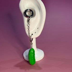 Può includere: Un dilatatore per orecchio color argento con una catena e un ciondolo a forma di cetriolo verde. Il dilatatore è esposto su un modello di orecchio bianco su uno sfondo viola. Il ciondolo a forma di cetriolo è di colore verde brillante.