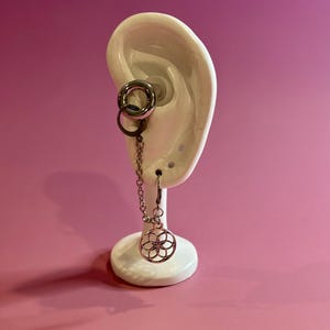 May include: A silver-colored ear gauge and chain earring set. The gauge is a circular design, and the earring features a chain that leads to a decorative, openwork floral pendant. The display is set against a pink background.