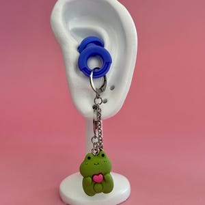 May include: A blue ear gauge with a silver chain holding a green frog charm with a pink heart. The frog charm is attached to a blue ear gauge, displayed on a white ear model against a pink backdrop.