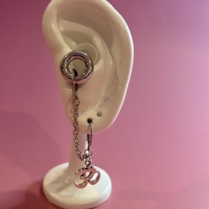May include: A silver ear gauge with a chain and an Om symbol charm. The gauge is in a white ear model, against a pink backdrop. The Om symbol is a spiritual icon. The gauge is a type of body modification.