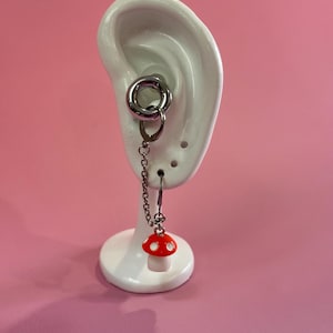 May include: A silver-toned earring set featuring a large ring and a chain with a red and white mushroom charm. The earring is displayed on a white ear model against a pink background.