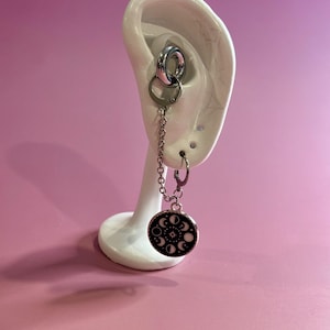 Può includere: Orecchini color argento a tema celeste. Gli orecchini presentano una catena che collega un orecchino a cerchio a un ciondolo circolare. Il ciondolo è nero con disegni bianchi di luna crescente e stelle. Gli orecchini sono esposti su un supporto bianco a forma di orecchio.