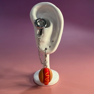Può includere: Un orecchino originale con un tunnel plug color argento e una catena. La catena sospende un ciondolo a forma di hot dog in miniatura. Il ciondolo ha un panino rosso, senape gialla e un panino marrone. L'orecchino è esposto su un supporto bianco a forma di orecchio.