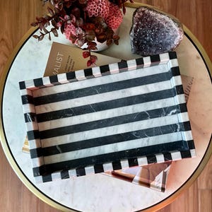 Puede incluir: Una bandeja decorativa rectangular con rayas blancas y negras. Descansa sobre una mesa con efecto mármol, junto a un jarrón con flores rosas y una geoda. La escena sugiere una elegante decoración del hogar.