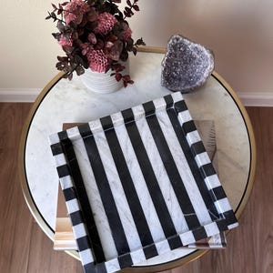 May include: A square tray featuring a bold pattern of alternating black and white vertical stripes. The tray has raised edges and a smooth, polished surface, showcasing a distinct contrast between the dark and light color bands throughout.