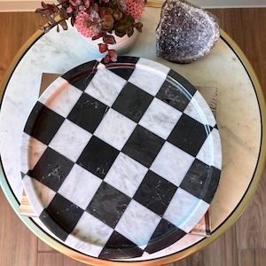 以下が含まれることがあります： 金色の縁が付いた丸い大理石のテーブルには、白黒の市松模様のトレイ、ピンクの花の配置、ジオードが置かれています。トレイのパターンは大胆なグラフィックデザインです。テーブルの表面は大理石模様です。